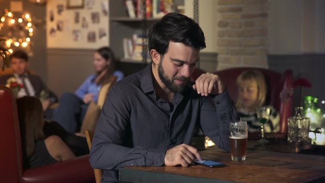 smiling man sitting at the bar with glass beer, receives a message on the phone