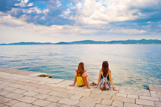 Two young girls with backpacks sitting on the stone near the sea shore, their hands leaning against stone pavement, looking at the hill range in the distance. Town of Zadar, Croatia
