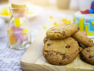 Close up of a chocolatechip cookies on wooden plate with colorful candy bottle on left side and sweet cake on back side