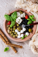 Mediterranean appetizer antipasti plate with green black olives, feta cheese, mozzarella, capers, pepper, basil with grissini bread sticks over beige concrete texture background. Top view with space