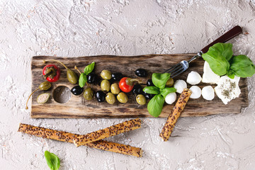 Mediterranean appetizer antipasti board with green black olives, feta cheese, mozzarella, capers, pepper, basil with grissini bread sticks over beige concrete texture background. Top view with space