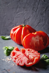 Whole and sliced organic tomatoes Coeur De Boeuf. Beefsteak tomato with pink salt and basil on blue gray metal texture background. Close up with space.