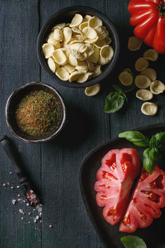 Homemade raw uncooked pasta with whole and sliced organic tomatoes Coeur De Boeuf, salt, seasoning, olive oil and basil over dark wooden background. Top view with space. Italian cuisine