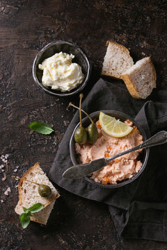 Black bowl of salmon pate with red caviar served with butter, sliced bread, capers, vintage knife and herbs on textile linen napkin over brown texture background. Top view.