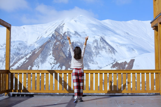 Happy healthy brunette woman in pajama back view stretching on balcony overlooking a mountain snow peak, sunny morning. Vacation in resort hotel. Simple lifestyle people in cozy relaxing apres ski