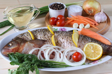 Fresh raw fish (ide) with slices of lemon lies on the dish before cooking among vegetables and greens.