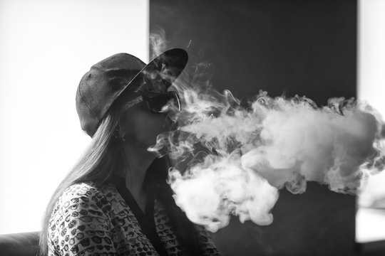 Vape. Young handsome white girl in sunglasses is admiting puffs of steam from the electronic cigarette. Vaping. Teenager. Black and white photo.
