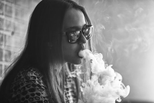 Vape. Young handsome white girl in sunglasses is admiting puffs of steam from the electronic cigarette. Vaping. Teenager. Black and white photo.