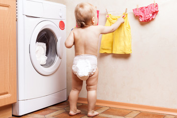 Baby stands near a washing machine and hanging wash clothes