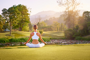 Yoga at park with view of the mountains, with sunlight. Young woman in lotus pose sitting on green grass. Concept of calm and meditation.