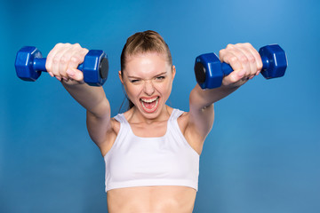 Fototapeta na wymiar aggressive young sportswoman training with dumbbells isolated on blue in studio