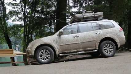 A dirty white SUV  with a roof luggage rack on top is parked at a campsite in the forest