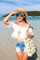 young cute happy girl on the beach, ocean. Dressed in denim shorts, top, straw hat, sunglasses, a bag with pineapple