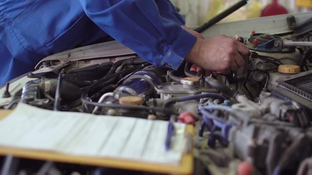 Car mechanic in a blue uniform fixing car under opened hood, diagnostics