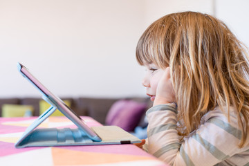 side view of three years old blonde child sitting indoor, with head over hands watching digital tablet on table with colorful tablecloth
