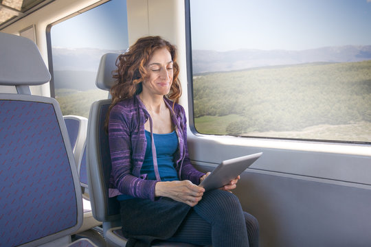 Red Hair Smiling Woman Dressed In Purple And Blue, Traveling By Train Sitting Reading Digital Tablet Or Ebook
