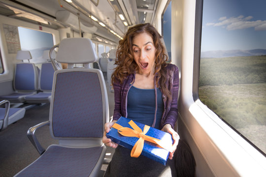red hair woman dressed in purple and blue, sitting traveling by train with gift box with bow in hands and amazed face expression
