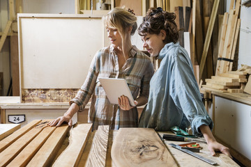 Two women choosing wood from a selection in a workshop, consulting on a tablet