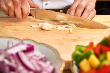 Man's hands cutting fresh garlic in the kitchen, preparing a meal for lunch. Paprika and onions in the foreground.