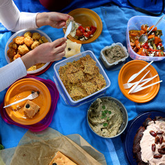 Unrecognizable person eating various picnic food: roasted vegetables salad, baba ghanoush, gluten-free crackers, foccacia bread, fried rice balls, gluten-free and sugarfree dates cake.
