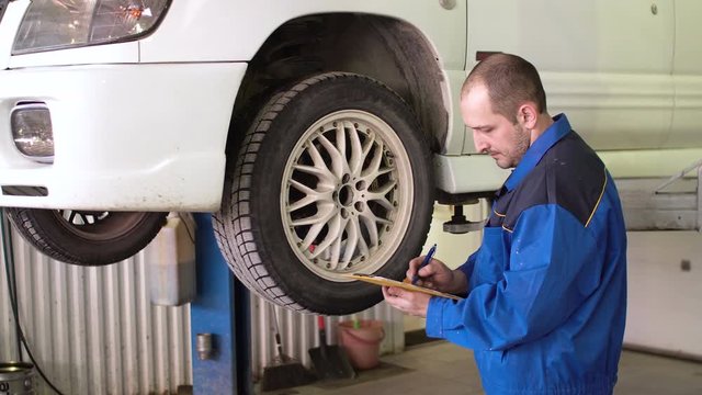 car mechanic is checking breaks and writing in notepad on lifted white car