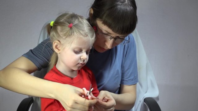 Mother in a blue turtleneck and glasses tonsured nails on the hands daughter in a red T-shirt using pink manicure scissors on a gray background.