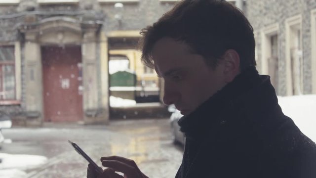 Portrait of young man in knitted scarf using smartphone on street under snowfall