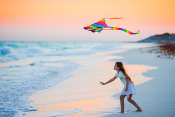 Little running girl with flying kite on tropical beach at sunset. Kids play on ocean shore. Child...