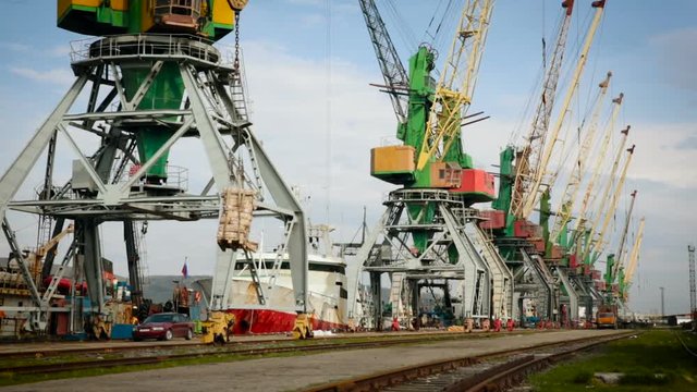Unloading Ships in Sea Port. Crane Moving Container With Load to Platform. Working Machines in Row. Boat Staying in Dock. Stone Path. Wide Shot