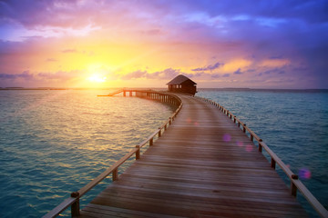 Obraz premium wooden road from the island to a hut over water on a sunset. Maldives...
