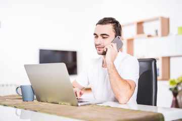 Handsome businessman is talking on the mobile phone and smiling while using a laptop in kitchen