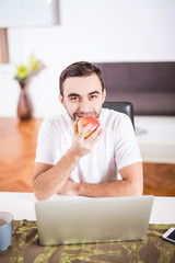 Handsome man eating apple and working with laptop at kitchen at home.