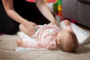 Mother changing diapers of a one year old baby girl daughter; child laying on the floor carpet in...