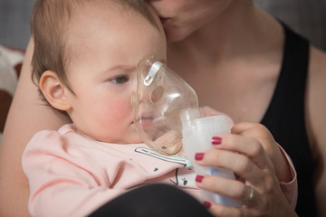 One year old baby girl inhaling from the inhaler, her mother holding her in the arms and comforting...