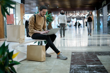 Obraz premium Student after buy clothes talk with friends in social network by laptop in shopping center. Man working during weekend walking. Immediate job for a freelance worker. Copy space for advertising sales.