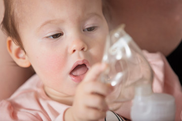 One year old baby girl holding the inhaler, her mother holding her in the arms and comforting the ill baby