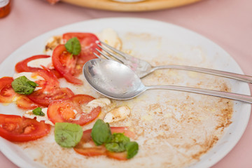 Tomato and mozzarella salad with basil leaves, sprinkled with pepper, olive oil and acceto balsamico - leftovers on the white plate; with spoon and fork