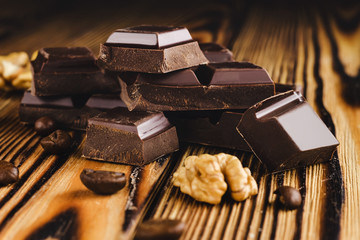 Pieces of bitter, dark chocolate, cubes on heap, walnut core and coffee bean on wooden background, close-up view