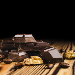 Pieces of bitter, dark chocolate, cubes on heap, walnut core and coffee bean on wooden background, close-up view