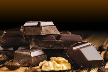 Pieces of bitter, dark chocolate, cubes on heap, walnut core and coffee bean on wooden background, close-up view