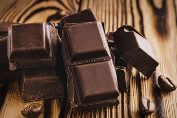 Pieces of bitter, dark chocolate, cubes on heap and coffee bean on wooden background, close-up view