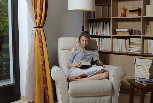 Young Woman Wearing Cozy Sweater,sitting In Comfortable Armchair, Enjoying Reading A Book At Home Library