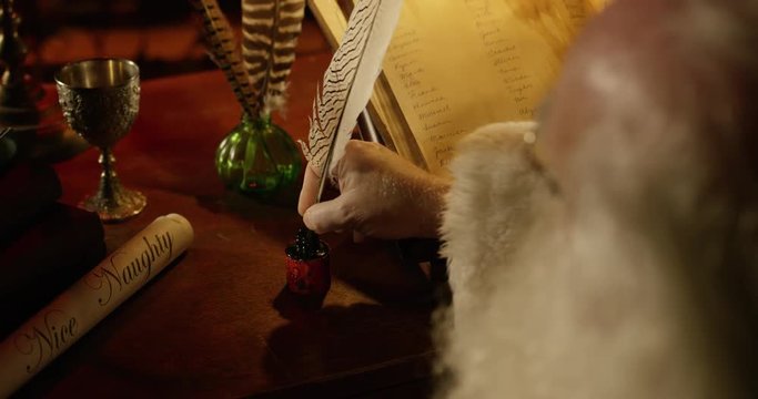 Santa Claus works on his naughty and nice list with a quill pen. Over shoulder.