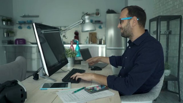 Man working on pc computer, taking break and drinking beverage by table at home
