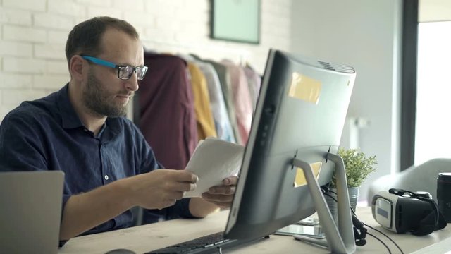 Unhappy, overwhelmed man working with documents and pc computer by table at home

