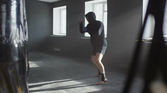 Side view of MMA fighter in hoodie shadow fighting and training knee kicks in empty gym lit by natural lighting coming from windows