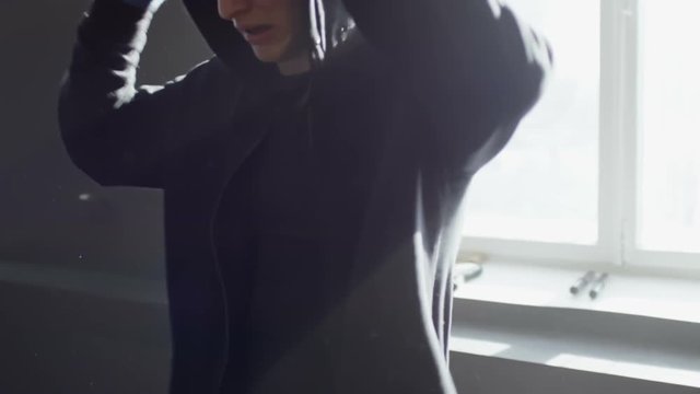 Slow motion of tired MMA fighter taking off hoodie and training punches before window in empty gym