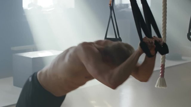 Handheld shot of muscly shirtless athlete doing push-ups on blast straps in empty gym