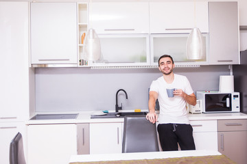 Fototapeta na wymiar Portrait of man standing in kitchen holding a cup of coffee looking away in thought. Relaxed young man having coffee at home in kitchen