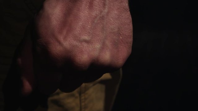 Hand of abusive male in yellow pants with swollen veins clenches fist in black semi dark room, close up shot